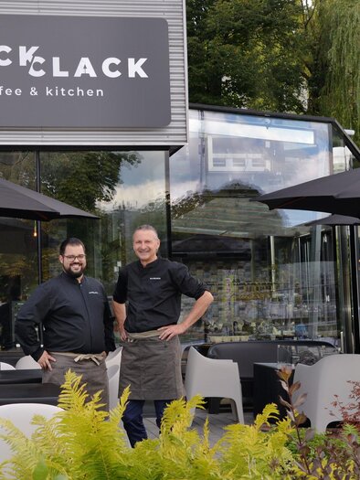 Zwei Männer stehen vor einem modernen Café mit dem Namen "CLACK coffee & kitchen". Der Außenbereich ist mit Tischen, Stühlen und Sonnenschirmen ausgestattet. | © Click Clack - Silvia Lambauer Fotografie