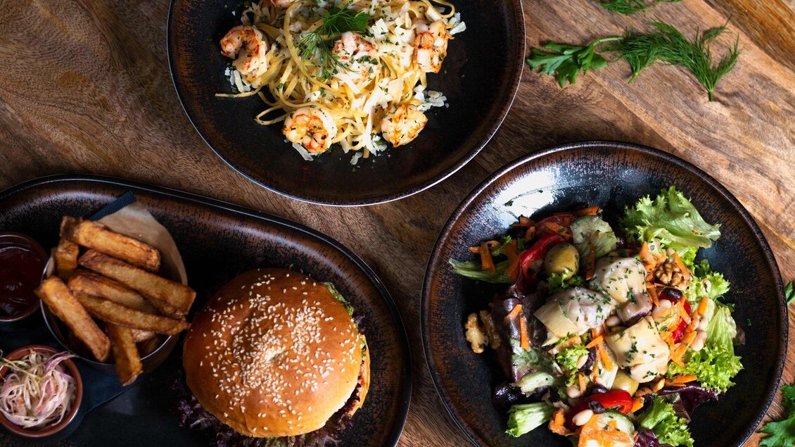 Ein köstliches Essen mit Pasta, Garnelen, Burger und frischem Salat. Dazu gibt es knusprige Pommes Frites auf einem Holzbrett. | © Click Clack - Ines Pötsch