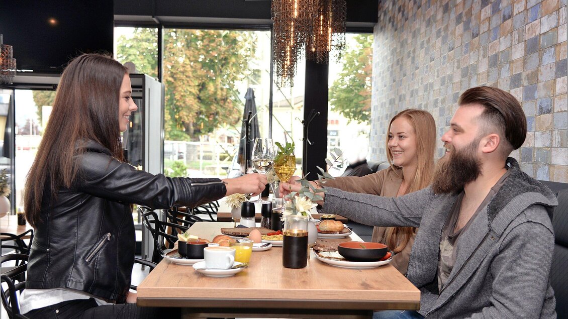 Drei Freundinnen stoßen in einem modernen Café mit Schaumweingläsern an. Auf dem Tisch sind verschiedene Speisen und Getränke angerichtet. | © Click Clack - Silvia Lambauer Fotografie