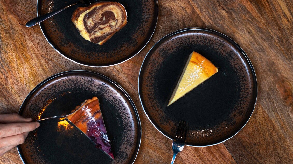 Drei Teller mit Desserts auf einer Holzoberfläche. Auf den Tellern befinden sich ein Stück Zopf, ein Stück Käsekuchen und ein Stück Fruchtkuchen. | © Click Clack - Ines Pötsch