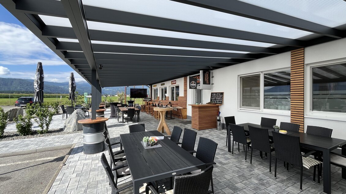 Eine gemütliche Terrasse mit Tischen und Stühlen unter einem modernen Überstand. Im Hintergrund ist ein schönes Panorama mit Bergen zu sehen. | © Peter Hopf