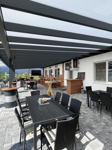 A cozy terrace with tables and chairs under a modern overhang. In the background, a beautiful panorama with mountains can be seen. | © Peter Hopf