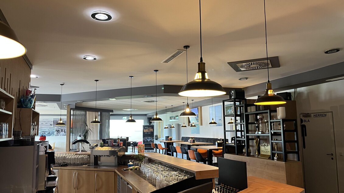 Ein modernes Restaurant mit holzverkleideter Bar und heller Beleuchtung. Tische und Stühle sind elegant angeordnet, und große Fenster bieten viel Tageslicht. | © Peter Hopf