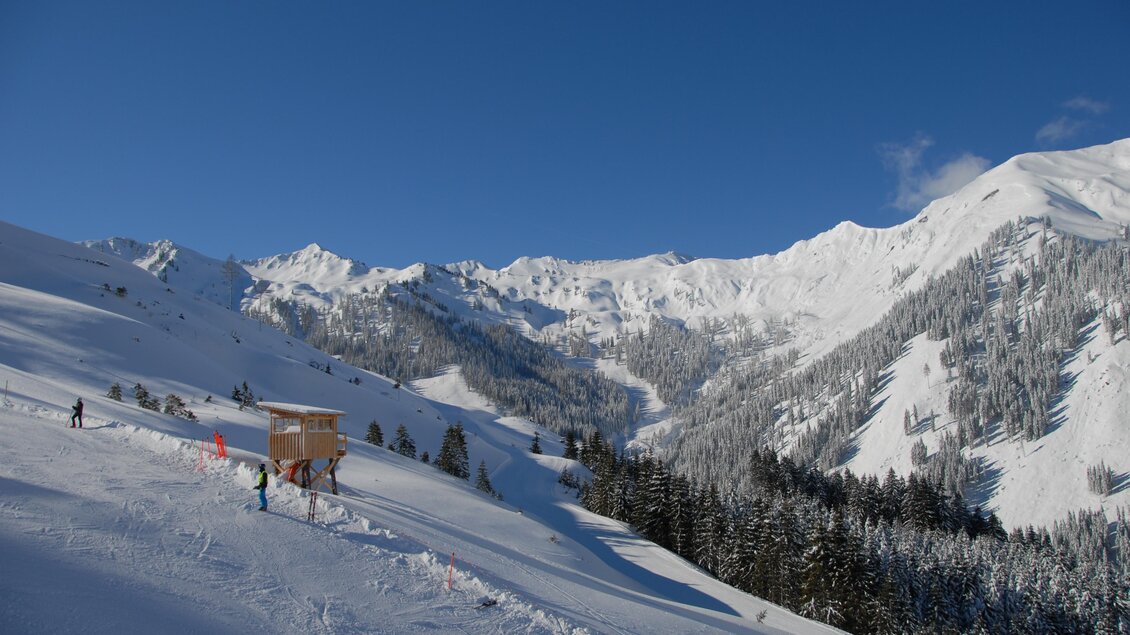 Eine verschneite Berglandschaft mit einem klaren blauen Himmel. Skifahrer genießen die Piste inmitten der schneebedeckten Berge. | © Riesneralm/Erwin Petz