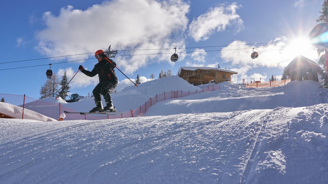Ein Snowboarder springt über einen verschneiten Hang. Im Hintergrund sind Skilifte und eine Berghütte zu sehen, bei klarem Himmel und Sonnenlicht.