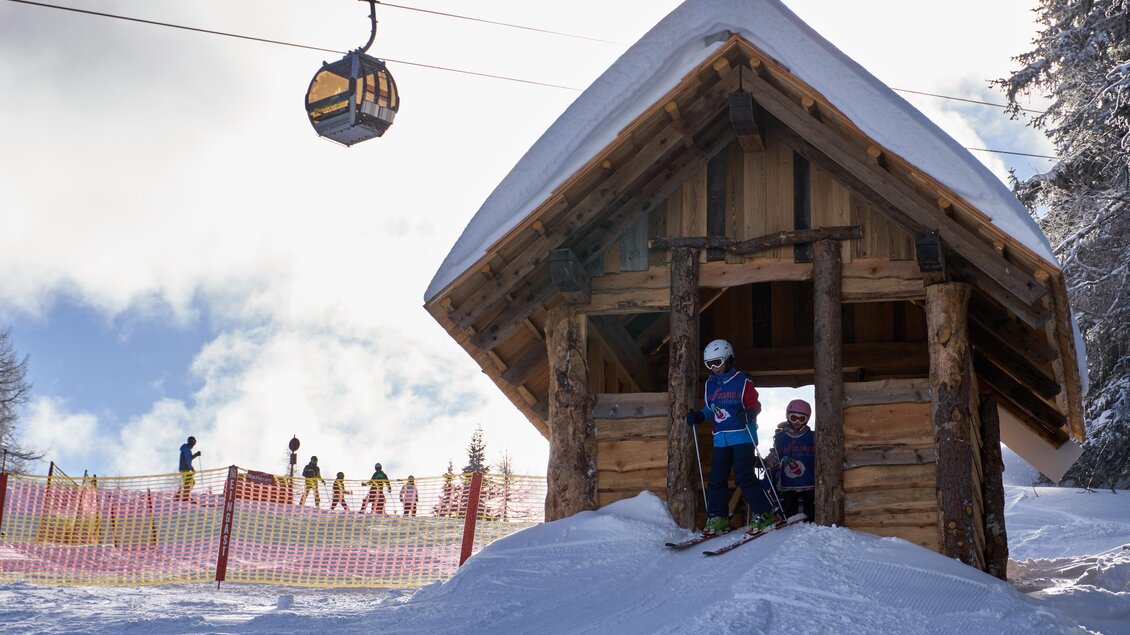 Eine Schneelandschaft mit einem kleinen Holzhaus und zwei Kindern in Skiausrüstung. Im Hintergrund sieht man Skifahrer und eine Gondel, die am Himmel schwebt.