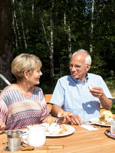 Ein älteres Paar sitzt an einem Holztisch im Freien und genießt gemeinsam eine Mahlzeit. Im Hintergrund sind Bäume und Grünpflanzen zu sehen. | © Region Graz - Harry Schiffer