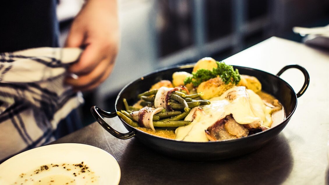 Ein köstliches Gericht in einer Pfanne mit Gemüse, Kartoffeln und Fleisch. Im Hintergrund ist ein Koch zu sehen, der das Essen anrichtet. | © TV Südsteiermark - Lupi Spuma