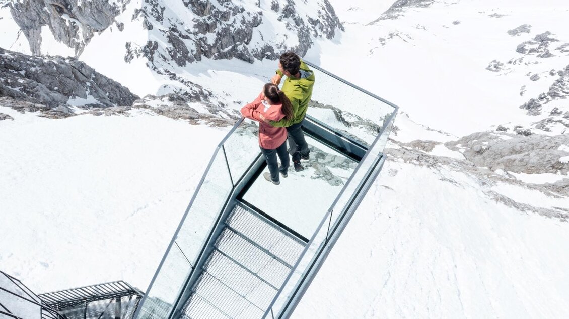 Ein Paar steht auf einer Glasplattform mit Blick auf schneebedeckte Berge. Die Umgebung ist kalt und majestätisch, umgeben von beeindruckender Berglandschaft. | © Harald Steiner