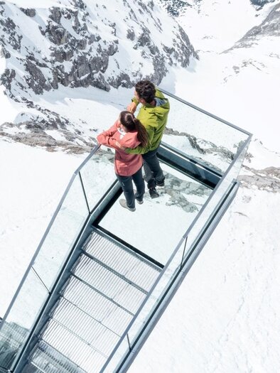 A couple stands on a glass platform overlooking snow-covered mountains. The surroundings are cold and majestic, surrounded by impressive mountain scenery. | © Harald Steiner