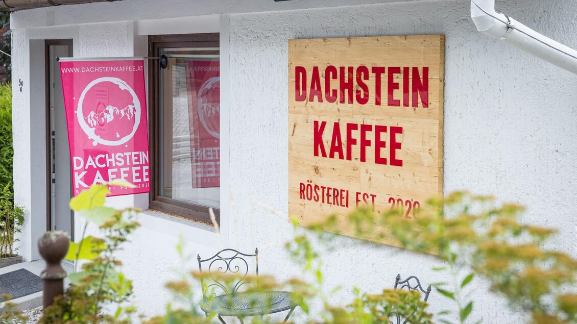 Ein gemütlicher Kaffee-Shop mit einem Holzschild, das "Dachstein Kaffee" zeigt. Vor dem Eingang stehen ein paar Stühle und Tische in einem einladenden Außenbereich. | © Wild und Team Fotoagentur GmbH