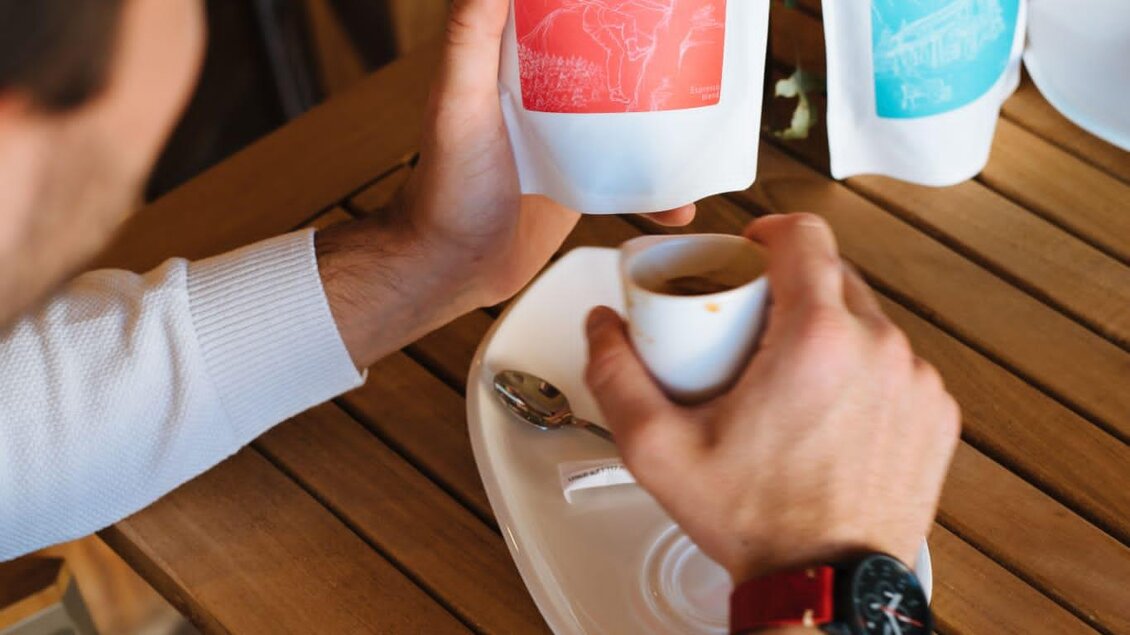 Eine Person hält eine Tasse Kaffee und eine Kaffeepackung auf einem Tisch. Im Hintergrund sind weitere Kaffeepackungen sichtbar. | © Lukas Bezila