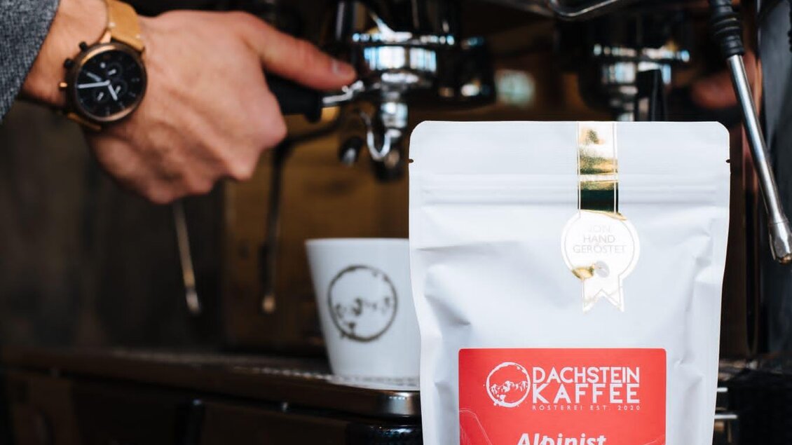 Eine Person bedient eine Espressomaschine, während ein Kaffeepaket mit dem Namen "Alpinist" im Vordergrund steht. Die Szene vermittelt eine gemütliche Kaffeeveraussicht. | © Lukas Bezila