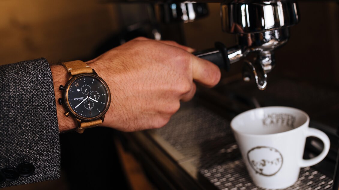 Eine Hand, die eine Espressomaschine bedient, trägt eine moderne Uhr. Vor ihr steht eine weiße Tasse mit dem Schriftzug "Kaffee". | © Lukas Bezila