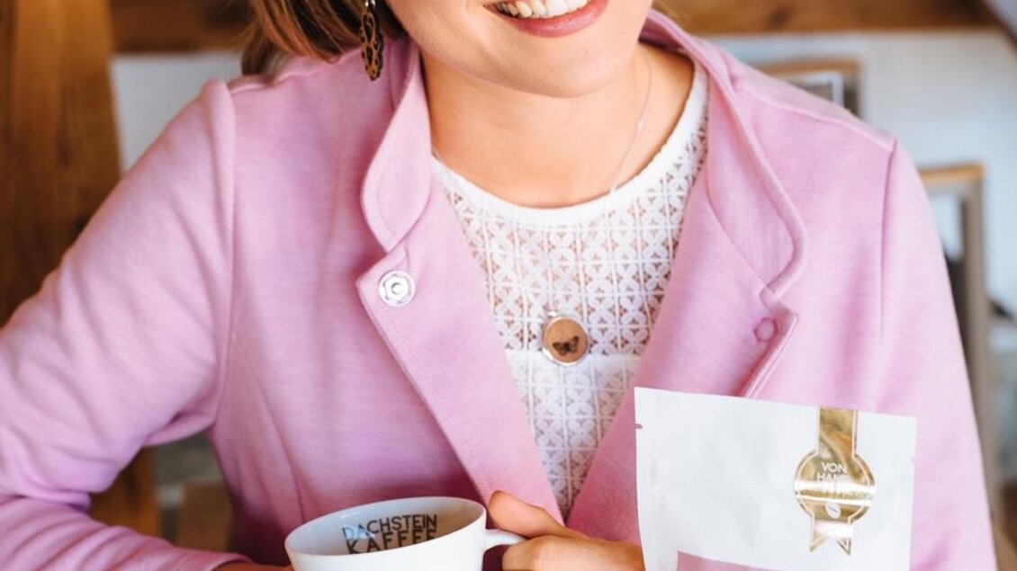 Eine lächelnde Frau hält eine Tasse Kaffee in der Hand. Neben ihr steht eine Packung Kaffee auf einem Tisch. | © Lukas Bezila