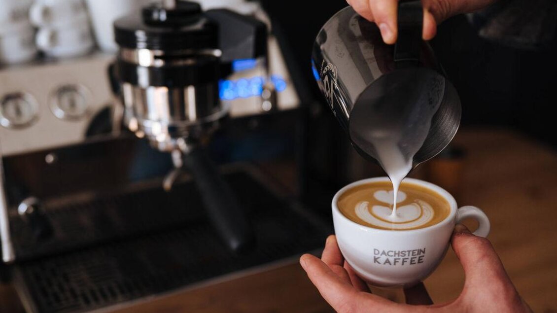 Eine Hand gießt Milch in eine Tasse Kaffee. Im Hintergrund ist eine Espressomaschine sichtbar. | © Lukas Bezila