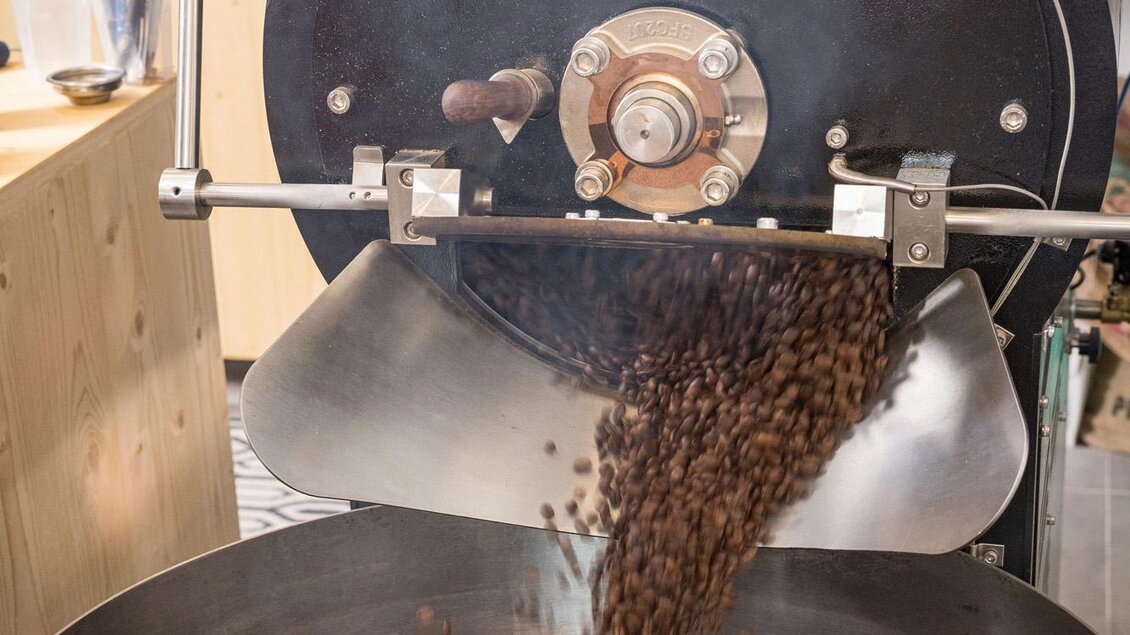 Eine Kaffeeröstmaschine, die frisch geröstete Kaffeebohnensorten in eine große Schüssel ablädt. Die Bohnen sind dunkelbraun und glänzend. | © Wild und Team Fotoagentur GmbH