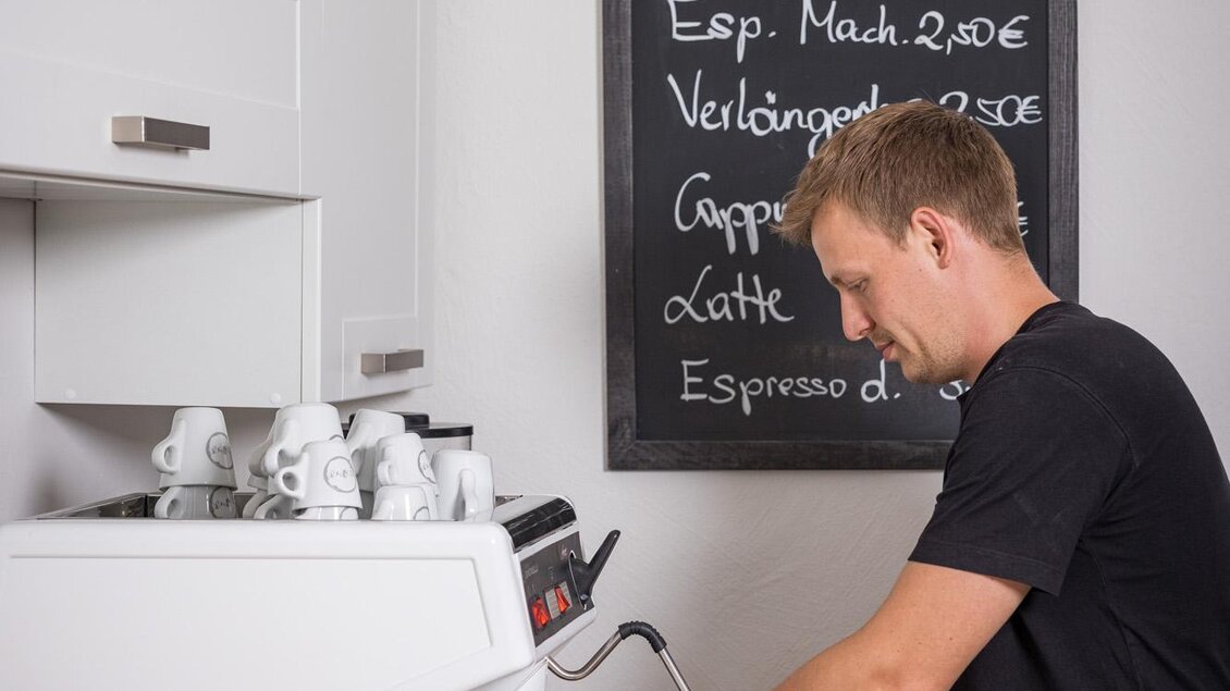 Ein Barista bereitet Kaffee an einer Espressomaschine in einer modernen Küche vor. Im Hintergrund ist eine Tafel mit den Getränkepreisen sichtbar. | © Wild und Team Fotoagentur GmbH
