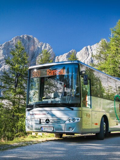 A bus drives through a picturesque mountain landscape. Surrounded by tall trees and impressive rocks, the scene offers a calm, nature-close atmosphere. | © René Eduard Perhab