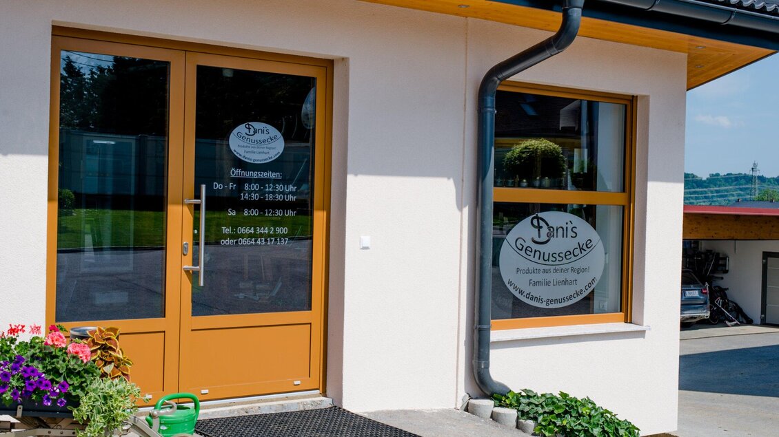 Ein einladender Eingangsbereich mit großen Fenstern und einer braunen Tür. Vor dem Eingang blühen bunte Blumen. | © TV Region Graz - Mias Photoart