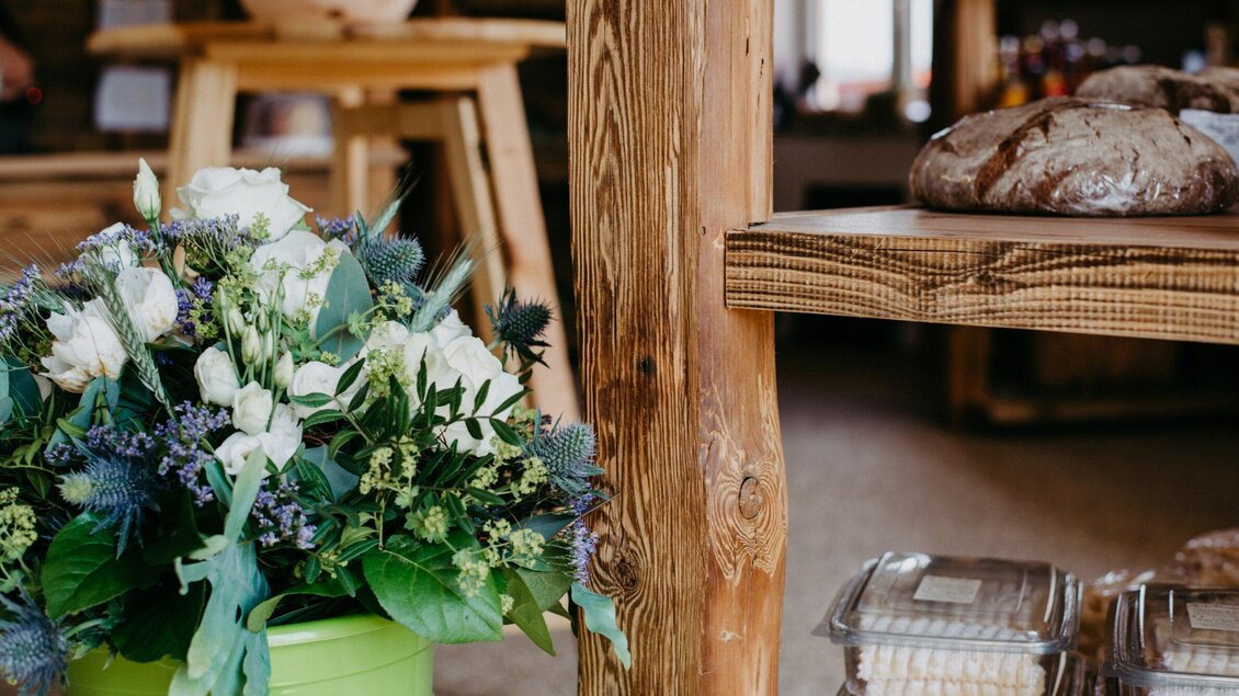 Ein hübscher Blumenstrauß in einem grünen Topf steht neben einem Holztisch. Auf dem Tisch liegen frisches Brot und andere Leckereien. | © TV Region Graz - Mias Photoart