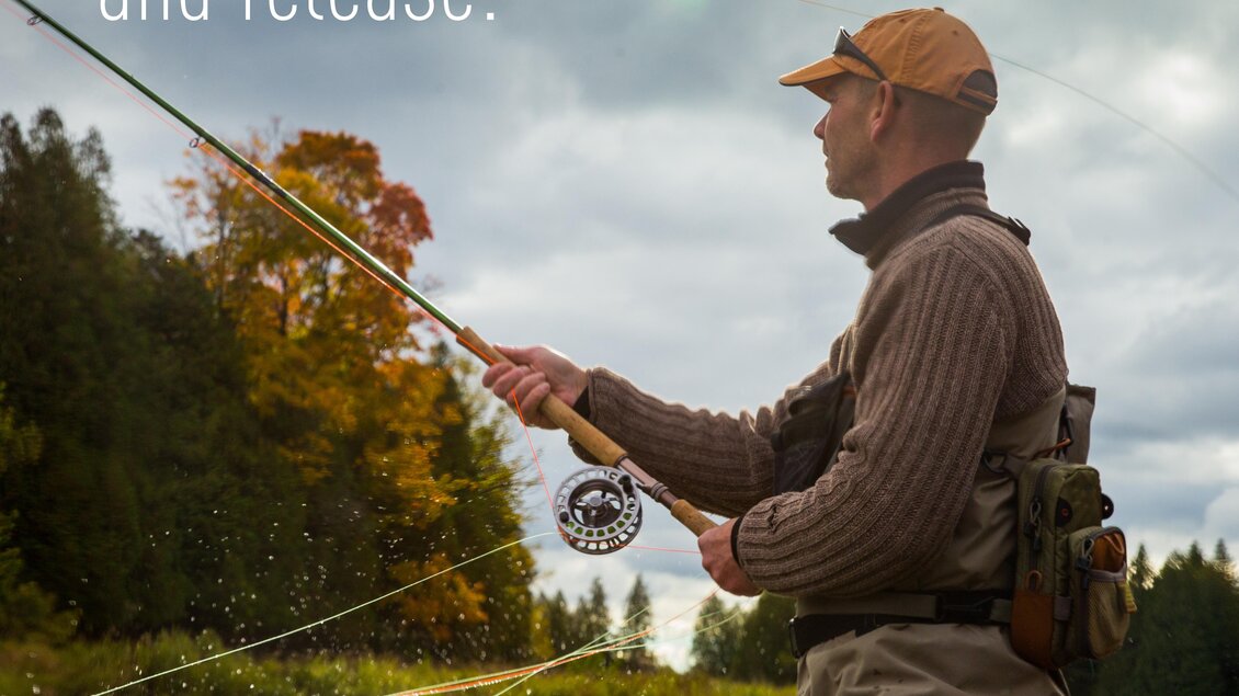Ein Angler steht im Wasser und fischt mit einer Angelrute. Im Hintergrund sind Bäume und ein bewölkter Himmel zu sehen.