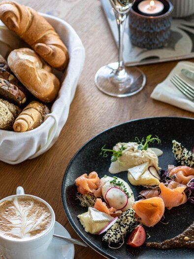 Ein reichhaltiges Frühstück mit verschiedenen Speisen, darunter frisches Brot, Lachs und Gemüse. Dazu gibt es Kaffee und ein Glas Wasser. | © Das Franz