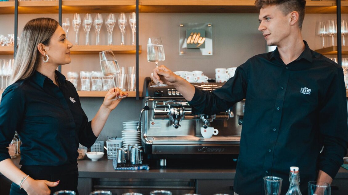 Hanna und Michael Paier stoßen in „Das Franz“ mit Gläsern an, Blick auf Kaffeemaschine und Regale. | © Das Franz