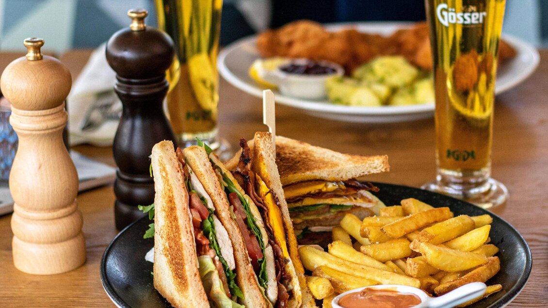 Ein leckeres Sandwich mit Kombinationen aus Fleisch, Käse und frischem Gemüse, serviert mit Pommes frites. Im Hintergrund stehen zwei Gläser Bier. | © Das Franz