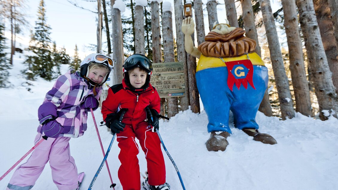 Zwei Kinder im Schnee stehen mit Skiausrüstung vor einer Holzfigur. Im Hintergrund sind schneebedeckte Bäume sichtbar. | © Tom Lamm