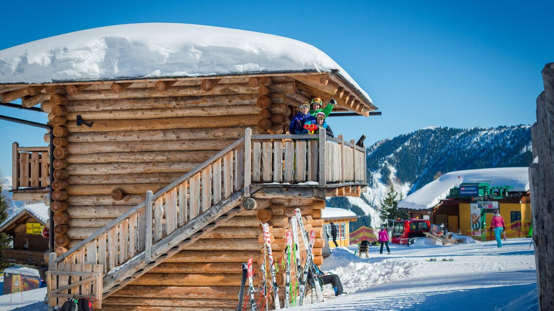 Eine charmante Skihütte aus Holz mit Schnee auf dem Dach. Im Vordergrund stehen fröhliche Skifahrer und Skiausrüstung auf einer verschneiten Piste. | © Christine Höflehner