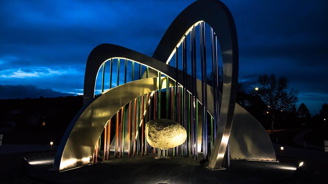 Eine moderne Skulptur mit leuchtenden Farben und geschwungenen Formen. Im Vordergrund befindet sich ein großer, runder Stein. | © TVB Bad Waltersdorf