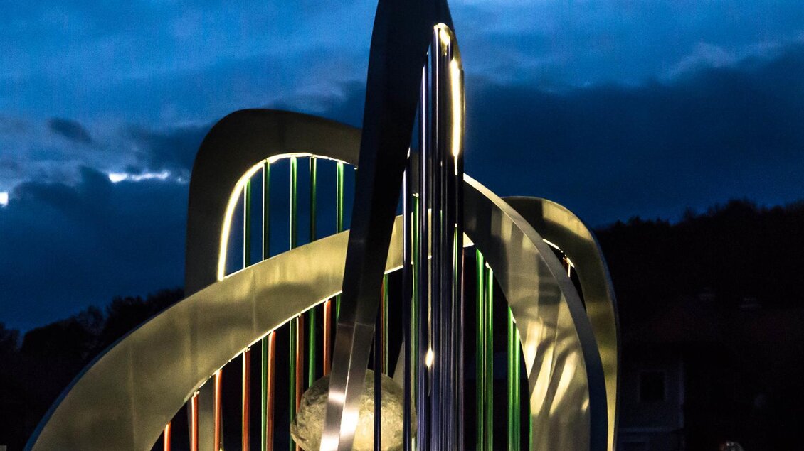 Eine künstlerische Skulptur mit bunten Lichtern vor einem dunklen Himmel. Die Formen sind geschwungen und modern gestaltet. | © TVB Bad Waltersdorf