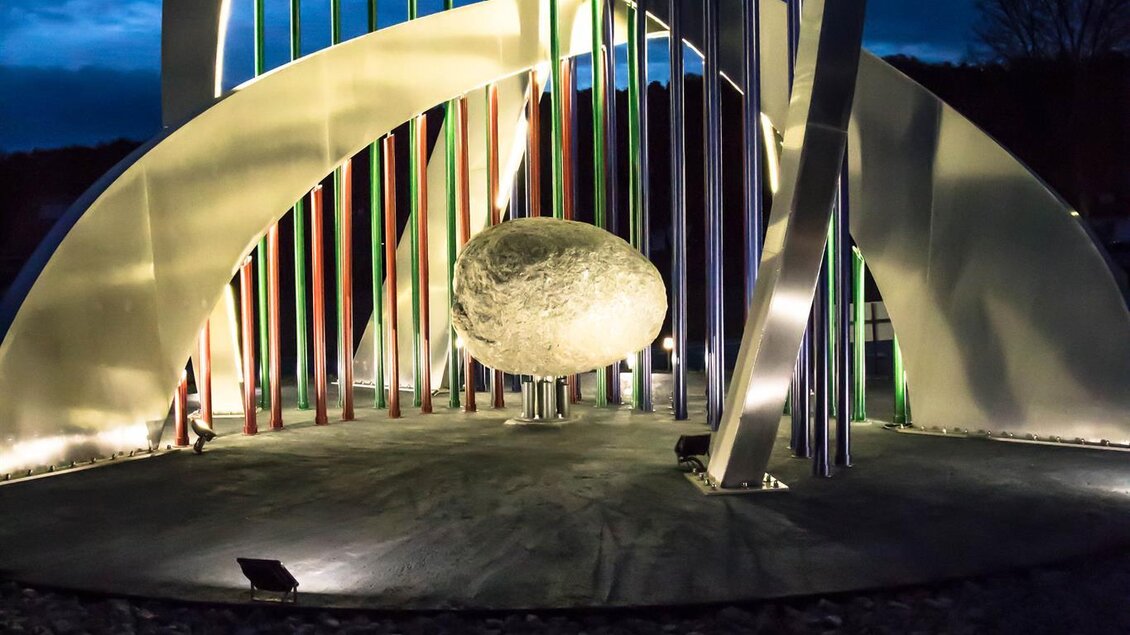 Eine moderne Skulptur mit bunten Lichtstrahlen und einem großen, runden Stein in der Mitte. Die Szene ist bei Nacht beleuchtet und hat eine futuristische Atmosphäre. | © TVB Bad Waltersdorf