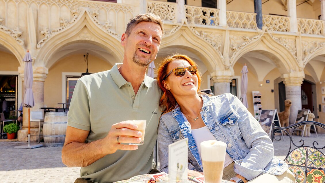Ein lächelndes Paar sitzt an einem Tisch in der Sonne und genießt Getränke. Im Hintergrund sind schöne architektonische Details zu sehen. | © Wolfgang Spekner
