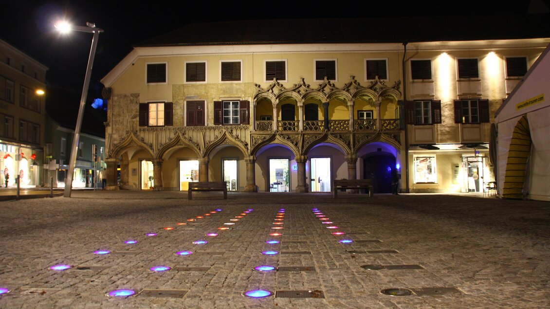 Ein Platz bei Nacht mit bunten Lichtern auf dem Boden und einem historischen Gebäude im Hintergrund. Die Atmosphäre ist ruhig und einladend. | © nicoleseiser.at
