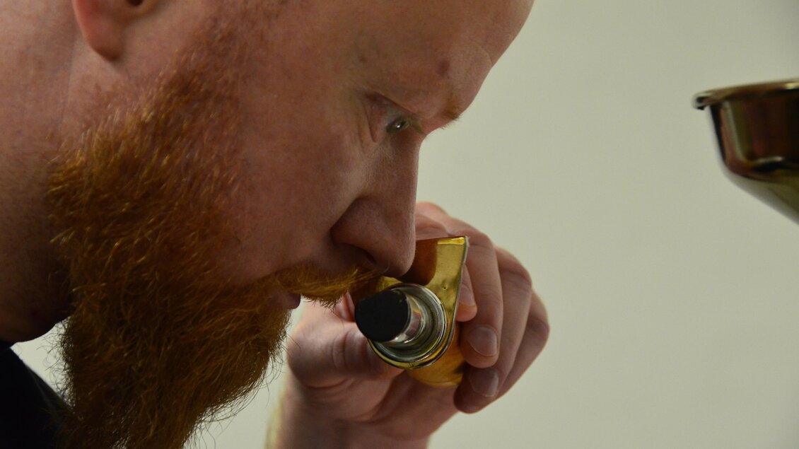 Ein Mann mit einem roten Bart beobachtet konzentriert durch ein spezielles Objektiv. Er arbeitet wahrscheinlich an einem kreativen oder handwerklichen Projekt.