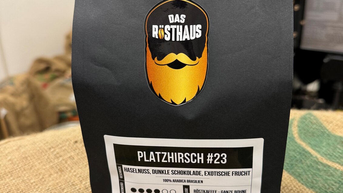 Eine schwarze Verpackung für Kaffee mit dem Namen "PLATZHIRSCH #23". Das Design enthält ein Bartlogo und Informationen über die Produktdetails.