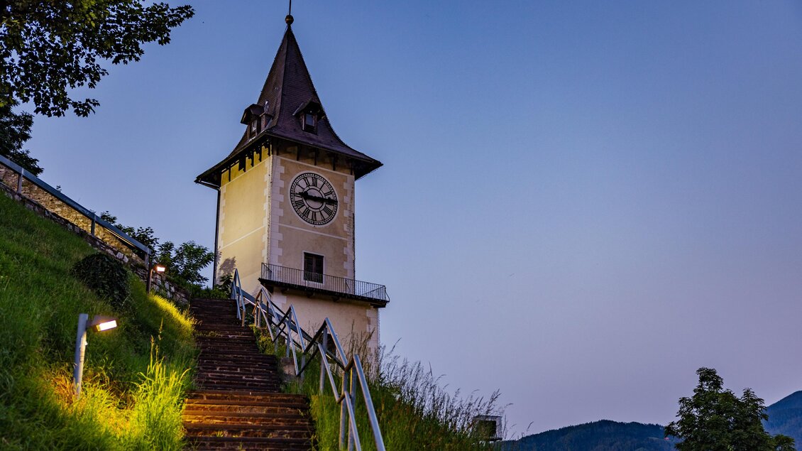 Ein historischer Uhrturm steht auf einem Hügel, umgeben von grüner Landschaft. Der Himmel ist in sanften Farben des Dämmerlichts gehalten. | © Wolfgang Speckner