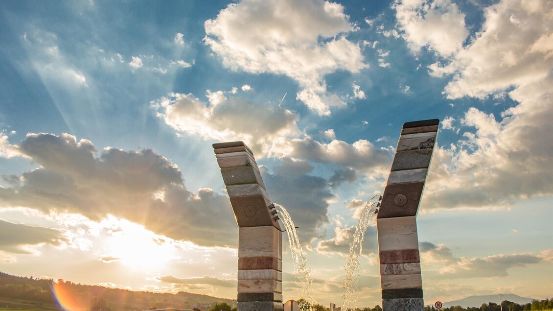Ein beeindruckendes Monument mit zwei großen, bogenförmigen Strukturen steht in einer grünen Landschaft. Im Hintergrund strahlt die Sonne durch eine Wolkendecke. | © Ingrid Jansky