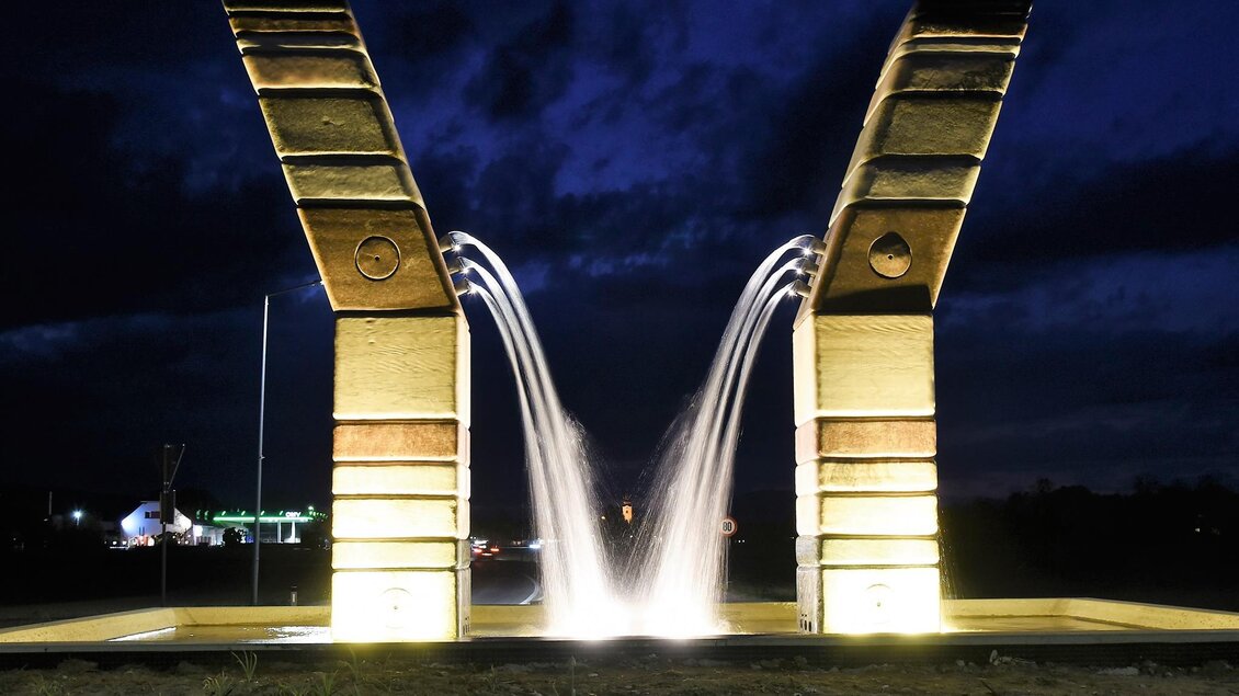 Ein modernes Wasserspiel mit zwei großen Bögen, das bei Nacht beleuchtet ist. Die Fontänen sprudeln zwischen den Bögen und erzeugen eine angenehme Atmosphäre. | © Lederer Josef