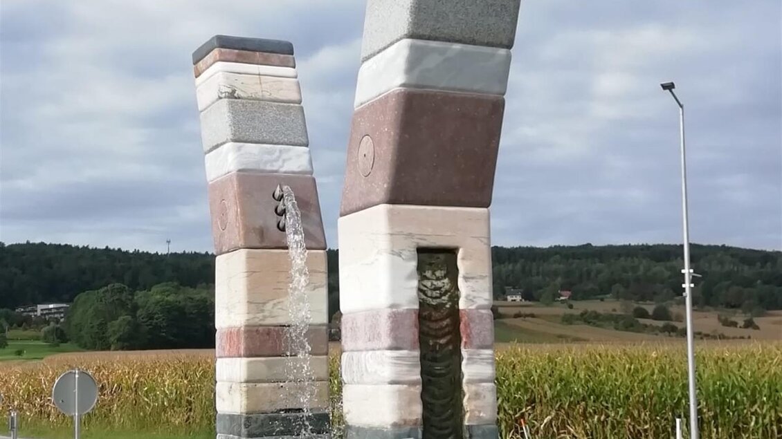 Eine moderne Steinsskulptur mit zwei schräg stehenden Säulen. Im Hintergrund sind Felder und ein bewölkter Himmel zu sehen. | © TVB Bad Waltersdorf