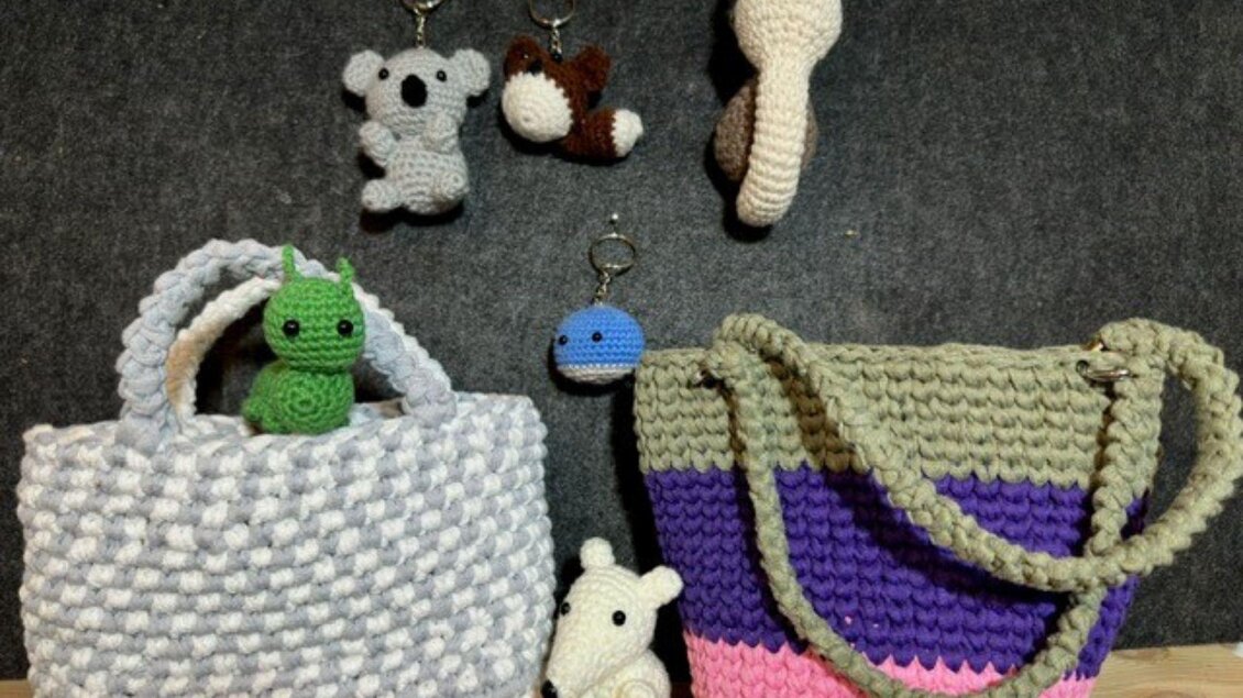 Hübsche, handgehäkelte Taschen in verschiedenen Farben stehen auf einem Tisch. Kleine, gehäkelte Tiere und Schlüsselanhänger sind ebenfalls zu sehen. | © Angelika Mikula-Hubacek