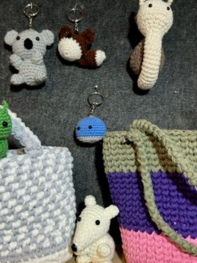 Hübsche, handgehäkelte Taschen in verschiedenen Farben stehen auf einem Tisch. Kleine, gehäkelte Tiere und Schlüsselanhänger sind ebenfalls zu sehen. | © Angelika Mikula-Hubacek