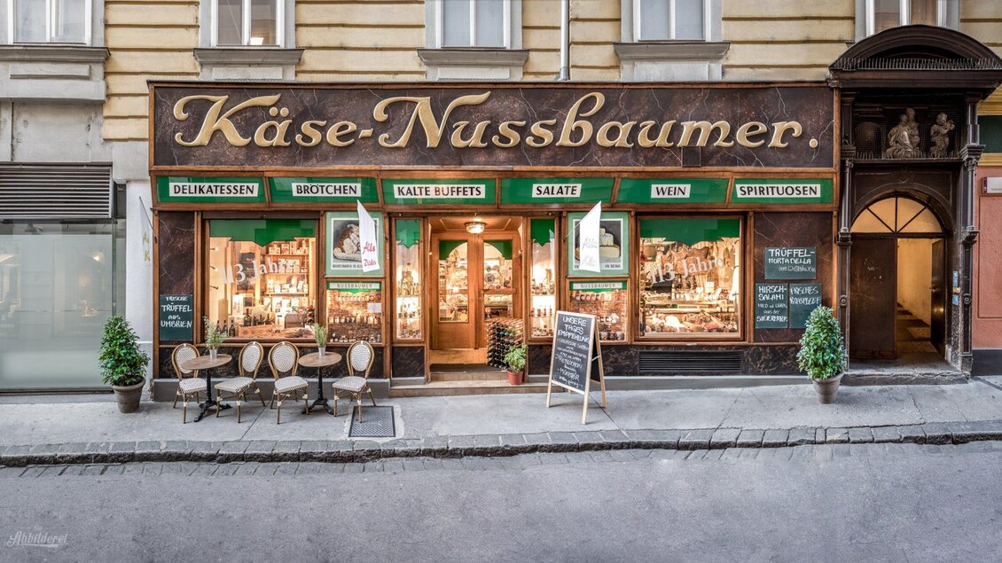 Ein einladendes Käsegeschäft mit dem Namen „Käse-Nussbaumer“. Es ist in einem historischen Gebäude mit großen Fenstern und einer gemütlichen Außensitzgelegenheit. | © Delikatessen Nussbaumer
