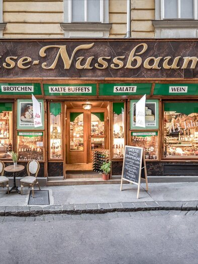 Ein einladendes Käsegeschäft mit dem Namen „Käse-Nussbaumer“. Es ist in einem historischen Gebäude mit großen Fenstern und einer gemütlichen Außensitzgelegenheit. | © Delikatessen Nussbaumer