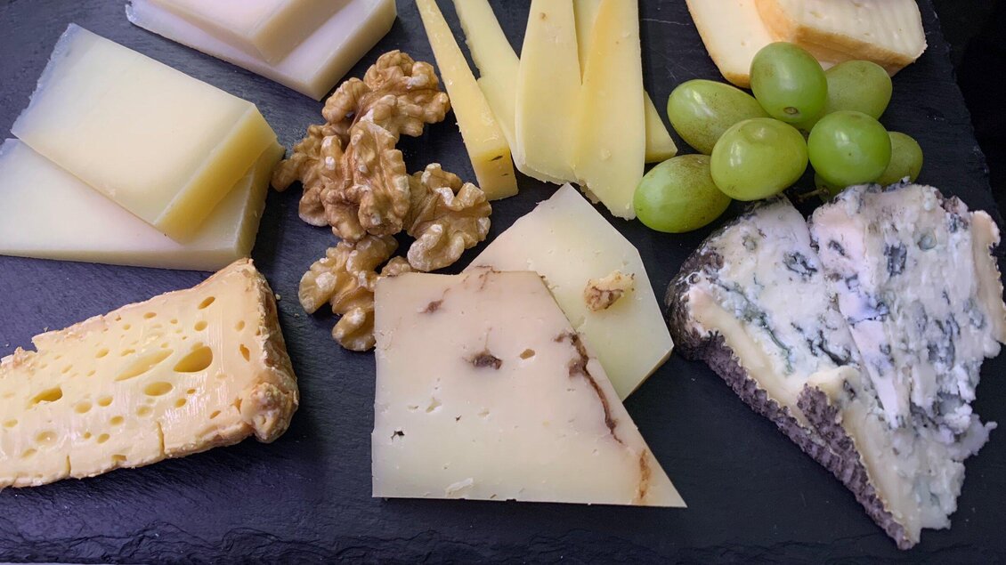 Eine Auswahl verschiedener Käsesorten mit Trauben und Walnüssen auf einem dunklen Brett. Die Käsesorten variieren in Farbe und Textur. | © Delikatessen Nussbaumer