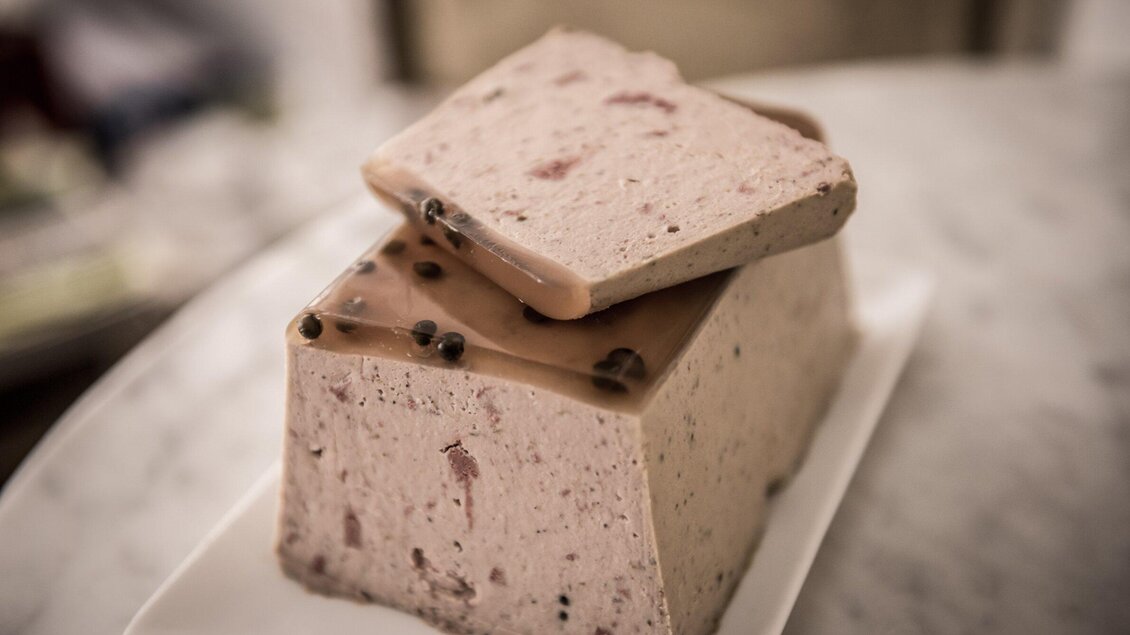 Ein Stück Pastete auf einem weißen Teller. Die Pastete ist hell mit Gewürzen und einer glasierten Oberfläche. | © Delikatessen Nussbaumer