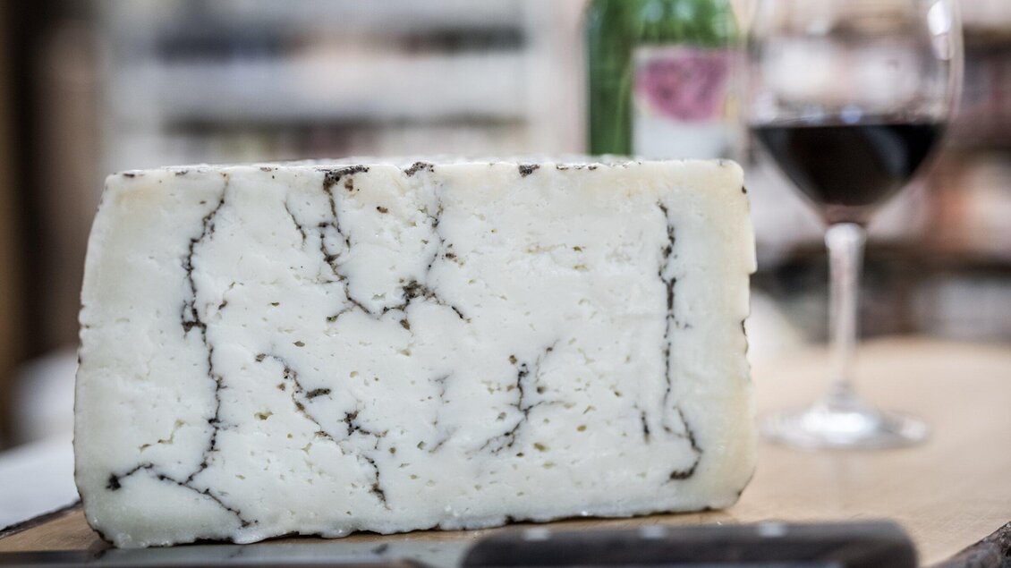 Ein Stück Käse mit schwarzen Adern liegt auf einem Holzbrett. Im Hintergrund sind ein Glas Rotwein und eine Flasche sichtbar. | © Delikatessen Nussbaumer