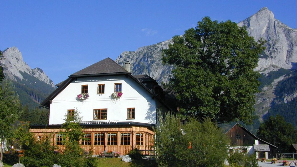 Ein malerisches Haus mit Holzveranda, umgeben von Bäumen und beeindruckenden Bergen. Der klare blaue Himmel ergänzt die idyllische Landschaft.
